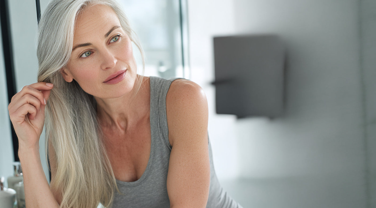 Woman with long, silver hair touching a strand in front of a mirror