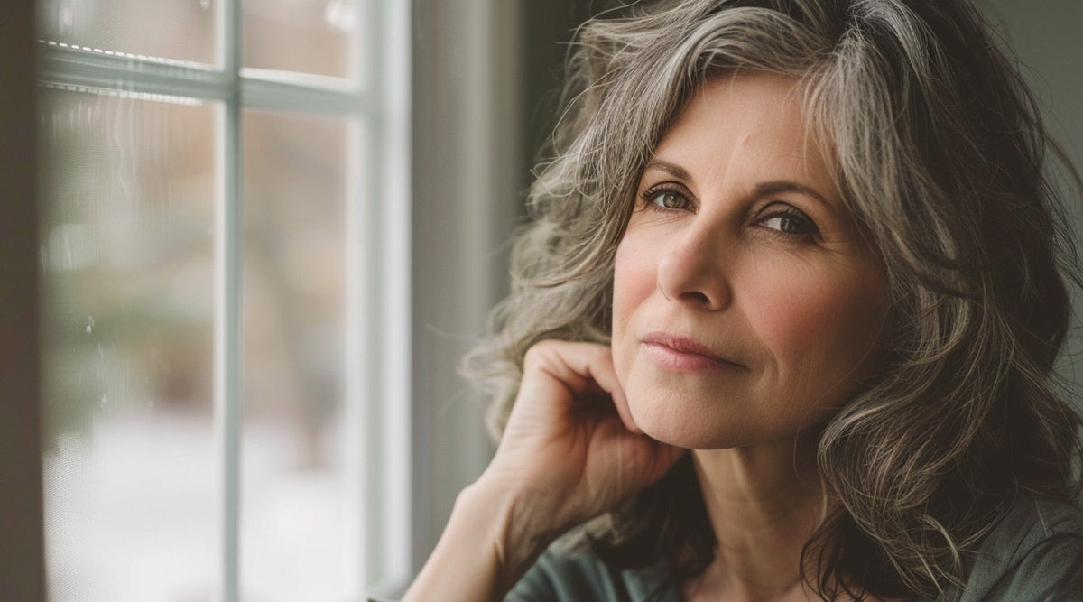 Woman with long, wavy gray hair resting her chin on her hand by a window