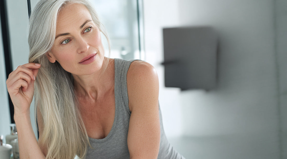 Woman with long, silver hair touching a strand in front of a mirror